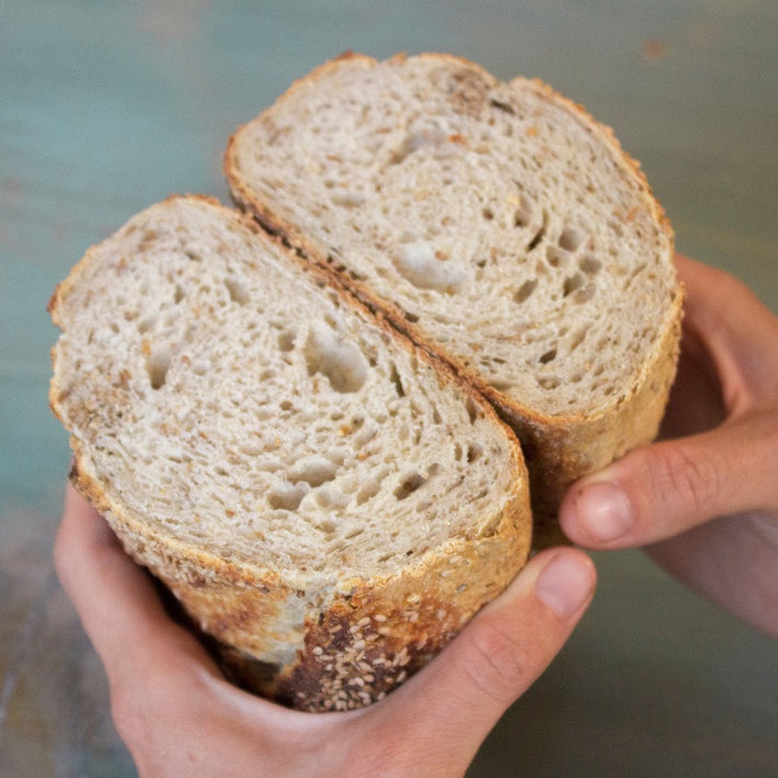Two hands holding a halved seeded sourdough loaf showing airy crumb and crust with sesame seeds.