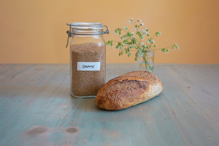 A jar of sesame seeds next to a sesame seed loaf on a wooden table with a small flower vase in the background.
