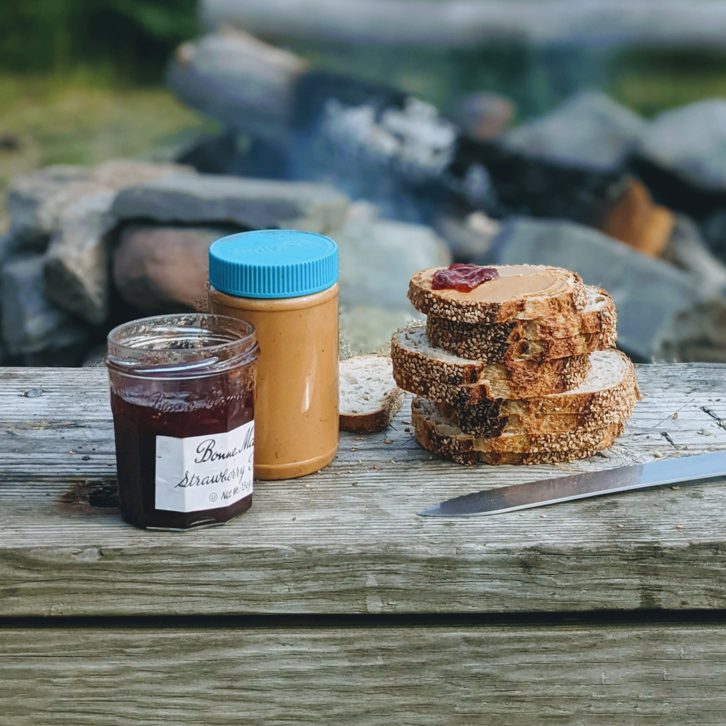 Jar of Bonne Maman Strawberry jam, peanut butter jar with blue lid, sesame bread slices and knife on a weathered bench.