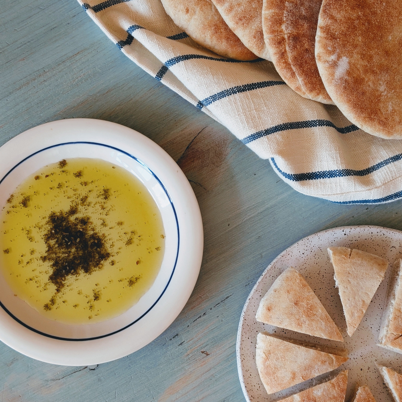Dish of olive oil with herbs beside whole and sliced pita on a blue wooden table with a striped cloth