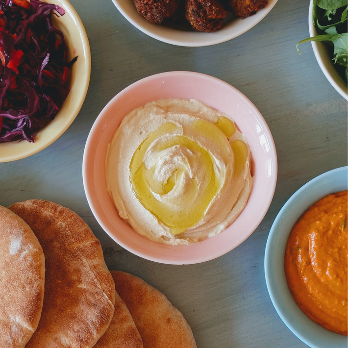 Pink bowl of hummus drizzled with olive oil, surrounded by pita, roasted red pepper dip, cabbage, and falafel on blue table
