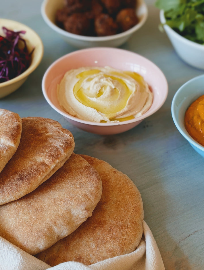 Close-up of soft pita bread stacked on a cloth, with hummus and salad bowls in the background.
