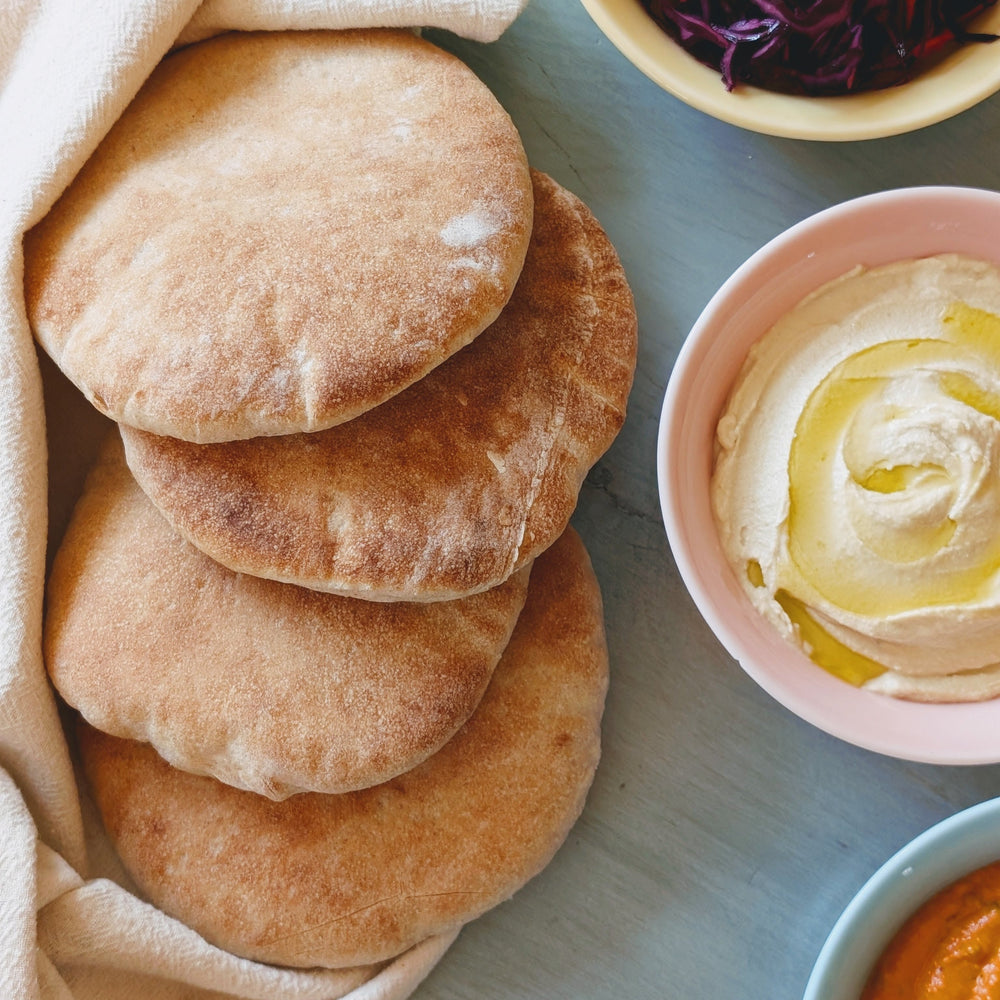 Stacked pita breads beside a bowl of whipped hummus drizzled with olive oil and two small bowls of dip