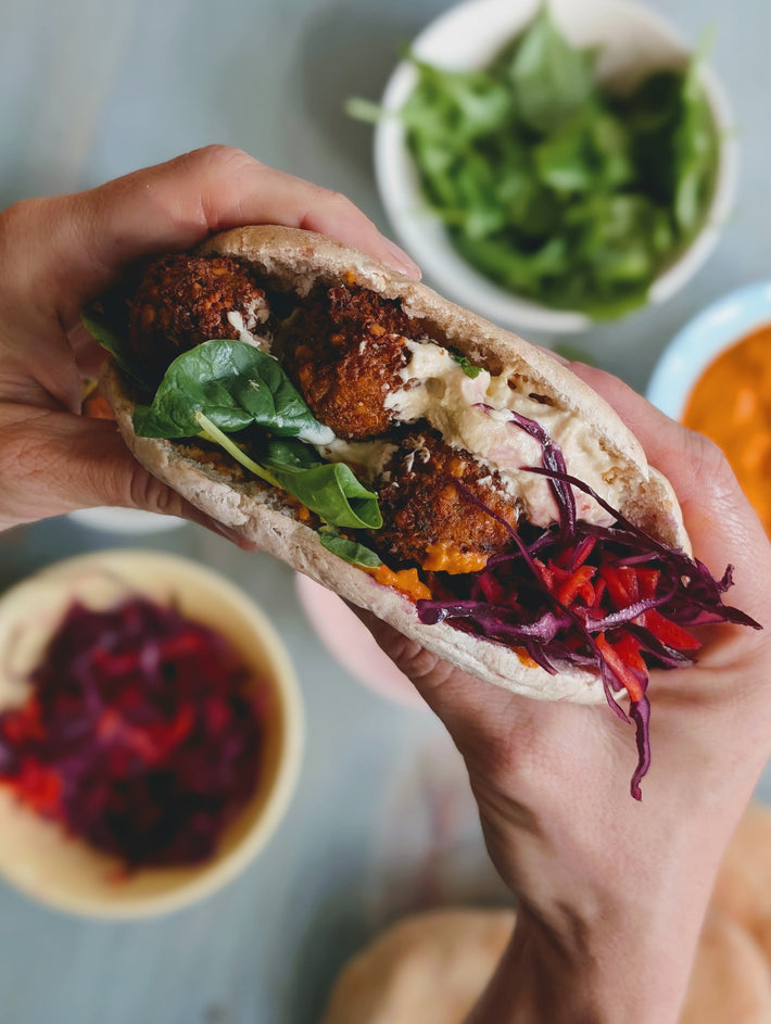 A person holding a pita filled with falafel, greens, and slaw, with bowls of salad and sauce in the background.