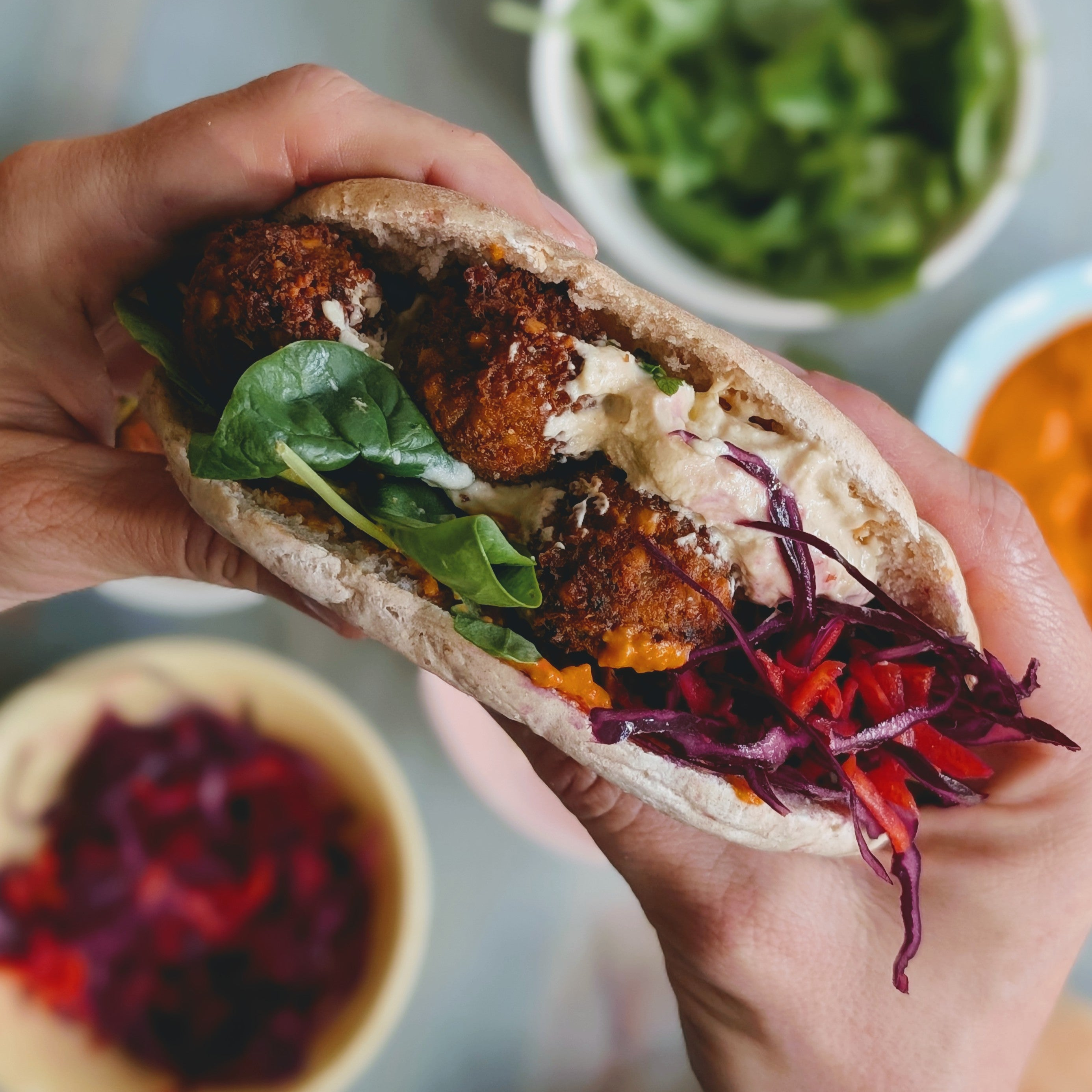 Pita stuffed with falafel, spinach, shredded red cabbage, and creamy sauce held by two hands over bowls.