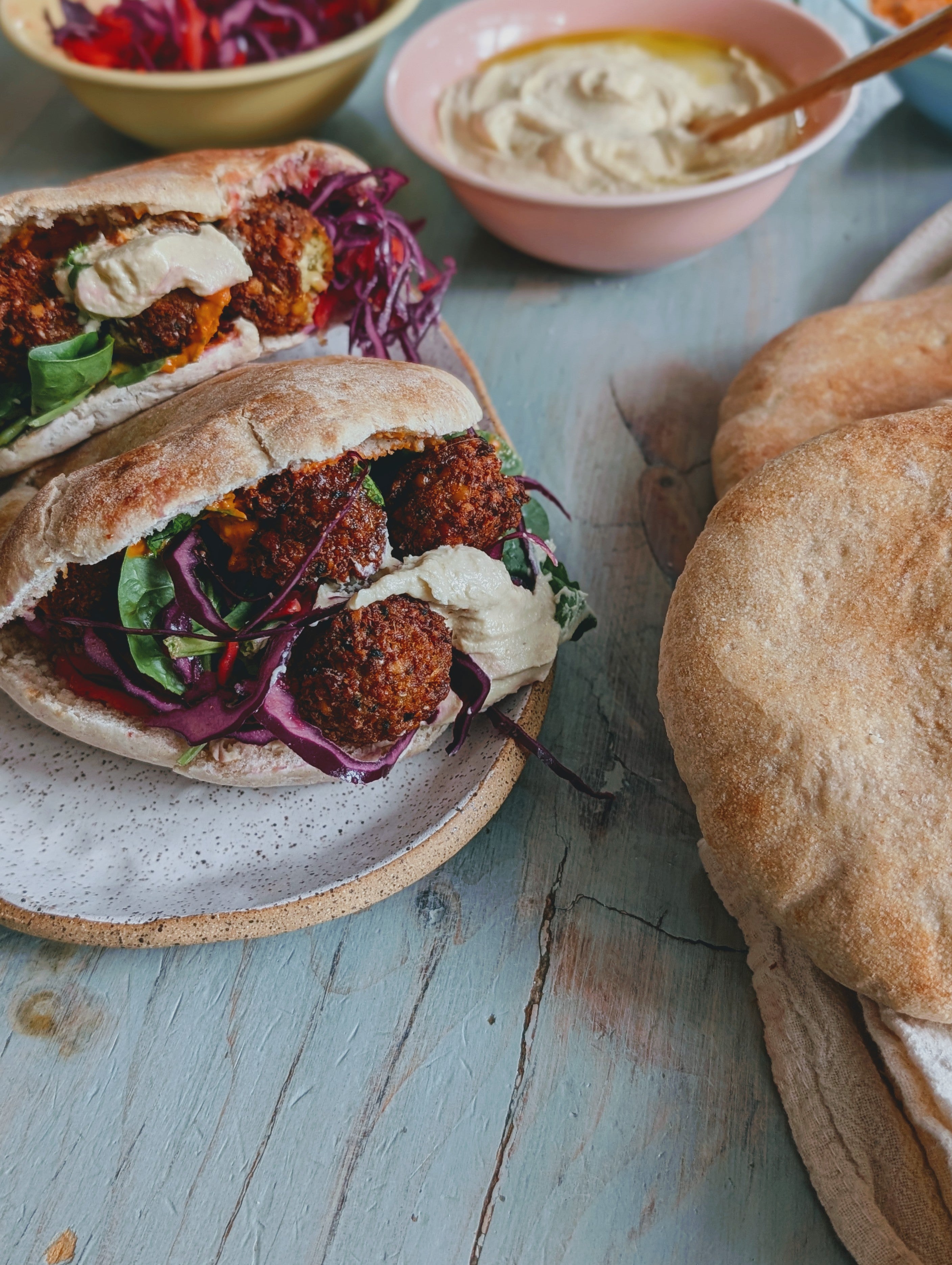 Two pita sandwiches filled with falafel, red cabbage, and greens, served with a side of hummus and pita bread on a rustic table.