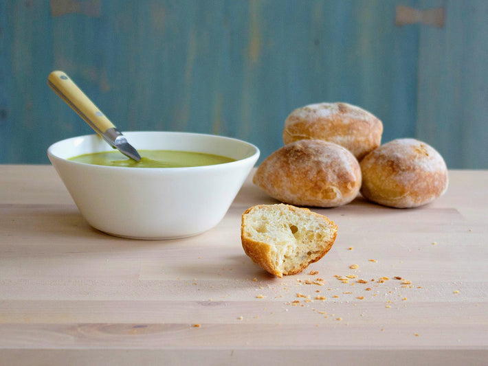 A bowl of green soup with a spoon, and three sourdough rolls on a wooden table.
