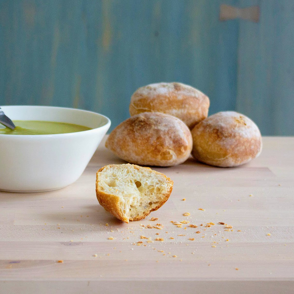 Crusty bread rolls with one torn roll and crumbs beside a bowl of soup on a wooden table.