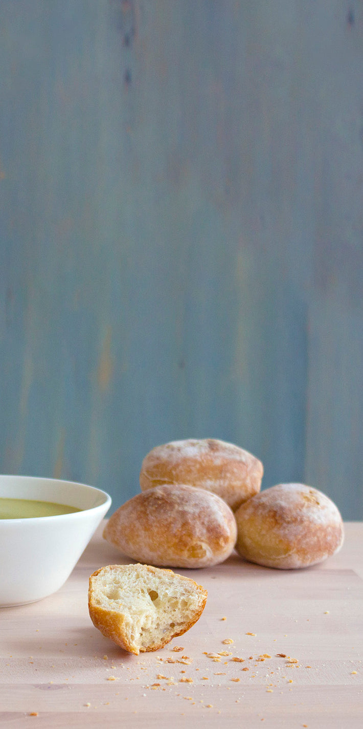 Small dusted round rolls—one torn with crumbs—next to a white bowl of olive oil on a light wooden surface.