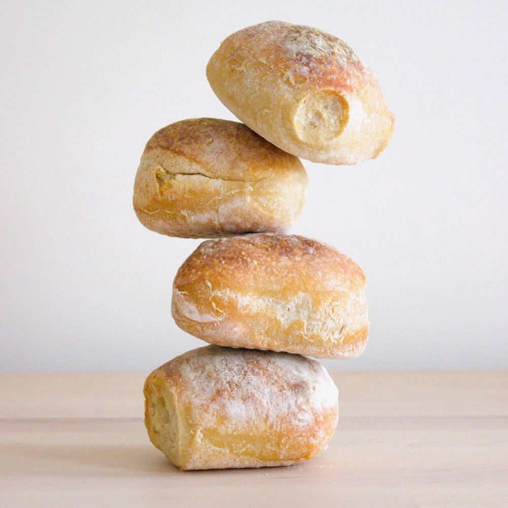 Four ciabatta rolls stacked vertically on a light wooden surface against a plain pale background.