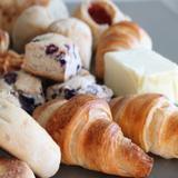 Close-up of Ready-To-Bake Catering Box contents showing golden-brown croissants, blueberry biscuits, and a block of butter on a baking sheet