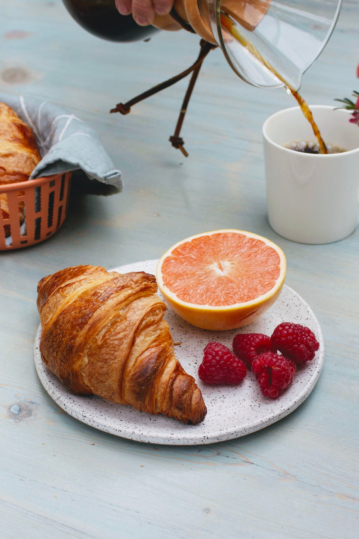 A croissant, raspberries, and a grapefruit on a plate with coffee being poured in the background.