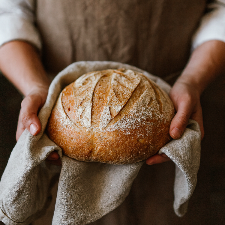 Hands holding a round scored sourdough loaf supported on a folded linen cloth