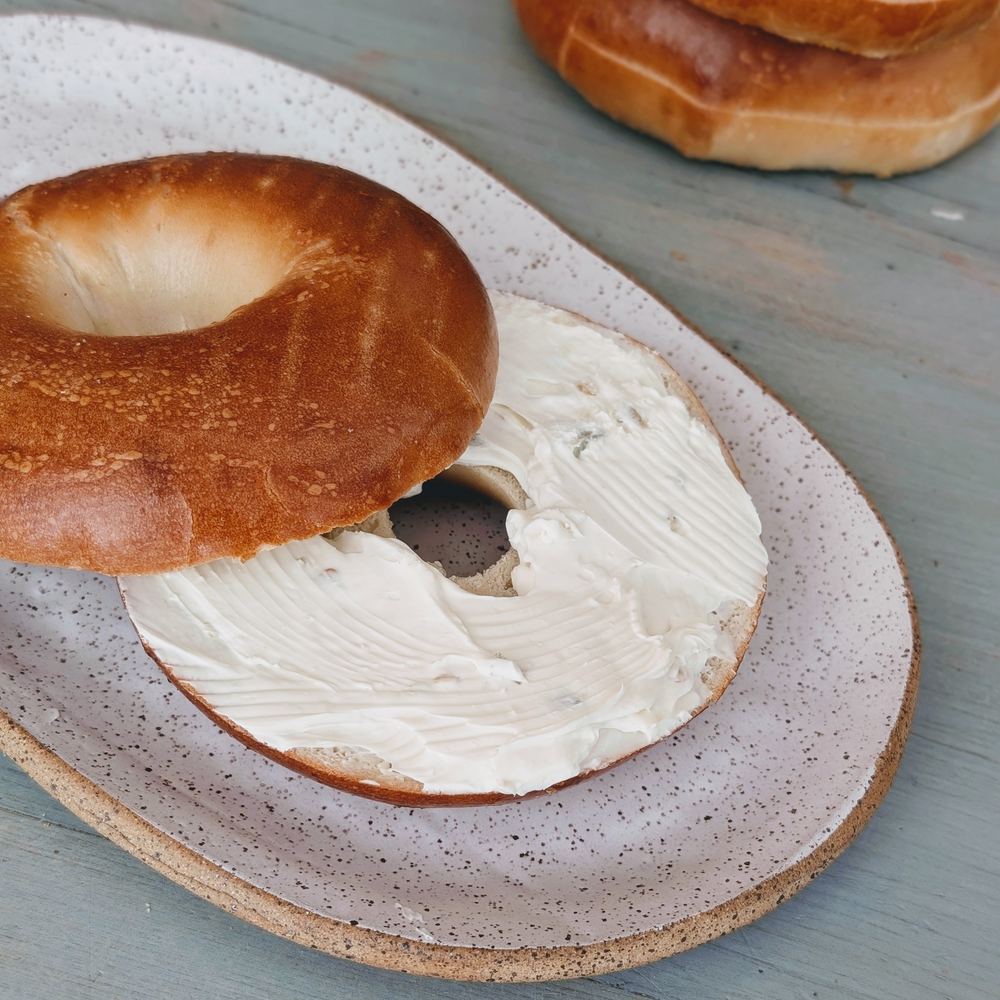 Toasted plain bagel, halved and spread with cream cheese, on a speckled ceramic plate atop a pale blue wooden table.