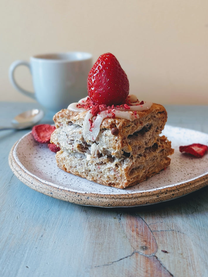 Close-up of strawberry white chocolate biscuit with white‑chocolate drizzle and fresh strawberry on top, on a speckled plate with a coffee mug in the background