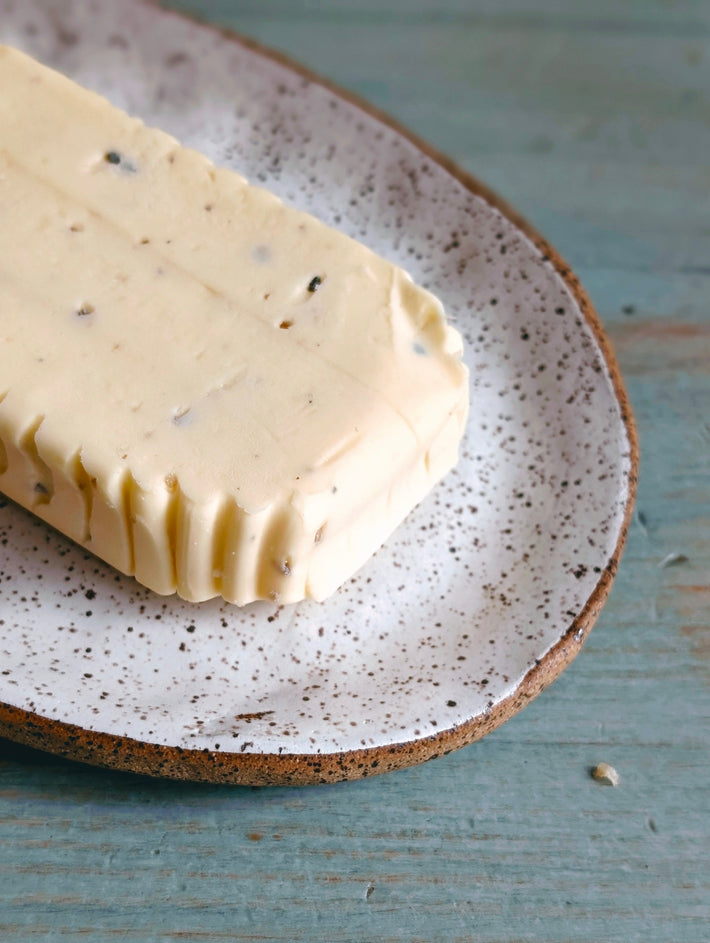 Stick of herbed compound butter with visible herbs on a speckled ceramic plate over a blue wooden surface.
