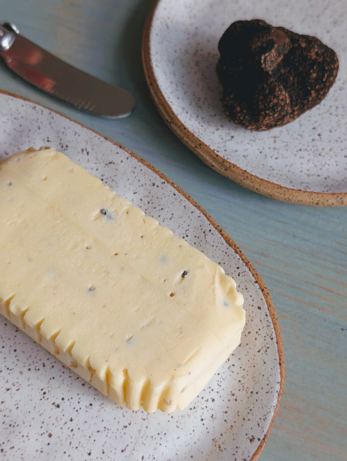 Block of truffle butter with visible truffle flecks on a speckled plate; whole black truffle and butter knife nearby.