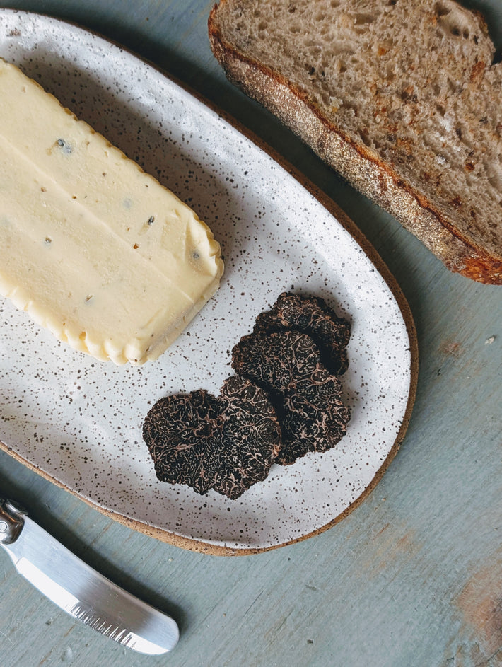 Speckled plate with a butter block and thinly sliced black truffles, bread slice at top and a butter knife at bottom.