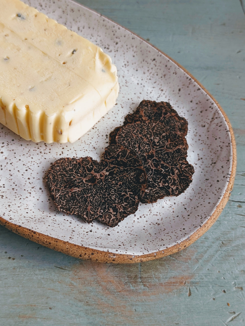 Block of herb butter and two thin black truffle slices on a speckled ceramic plate.