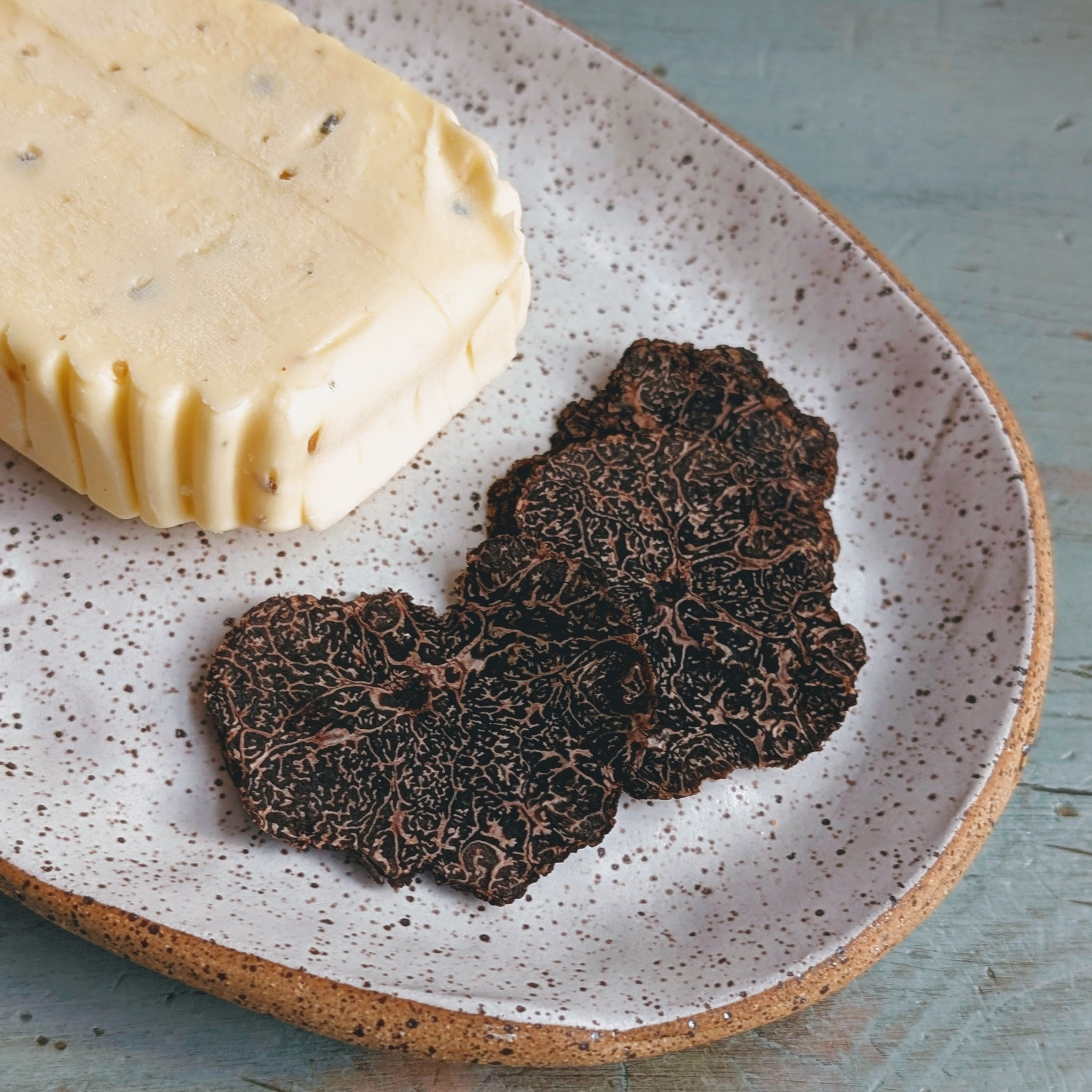Round pat of softened butter with two thin black truffle slices on a speckled ceramic plate.