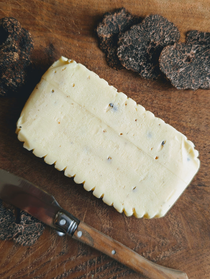 Rectangular slab of truffle butter with black truffle flecks on a wooden board, sliced truffles and a knife.