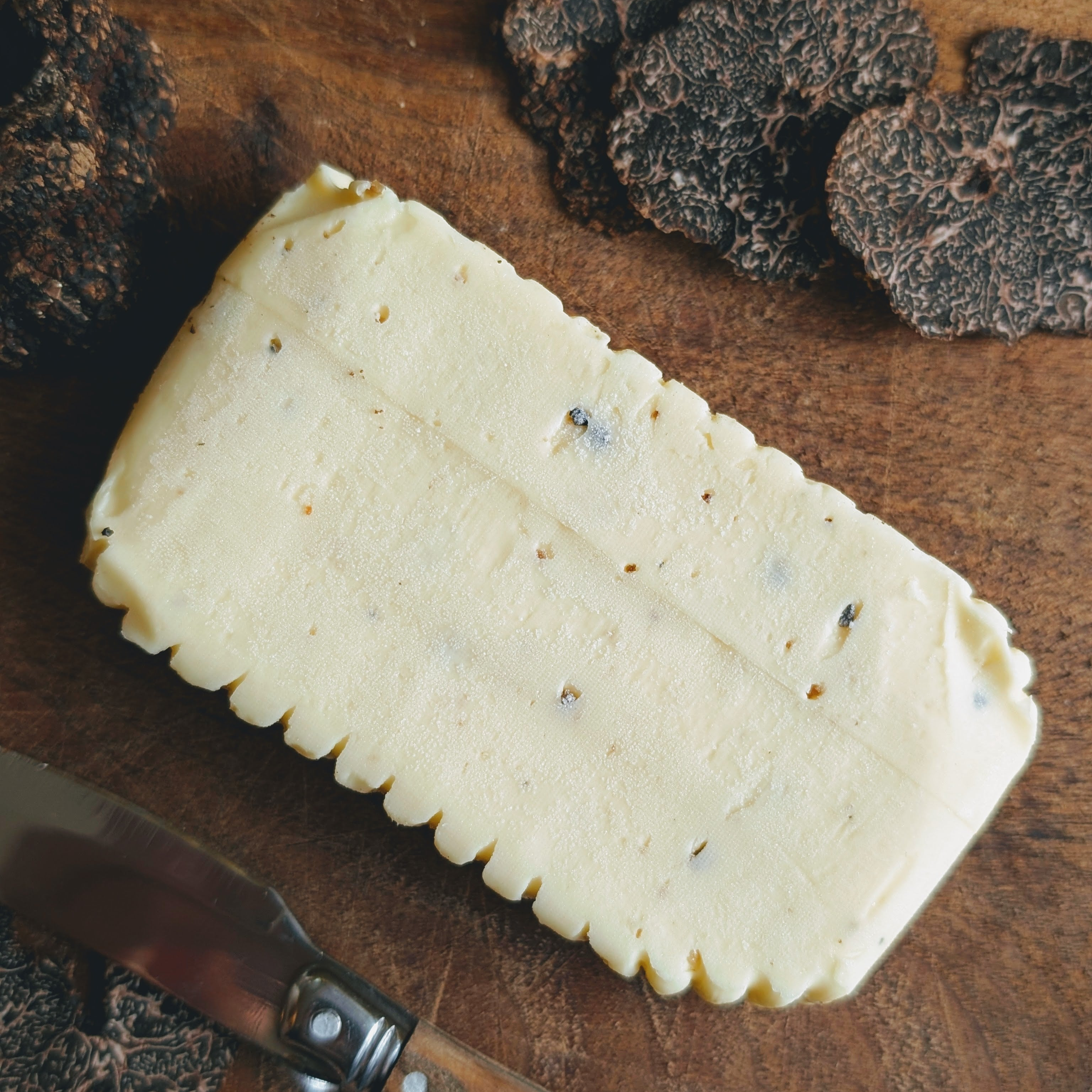 Rectangular slab of truffle butter on a wooden cutting board with sliced black truffles and a butter knife
