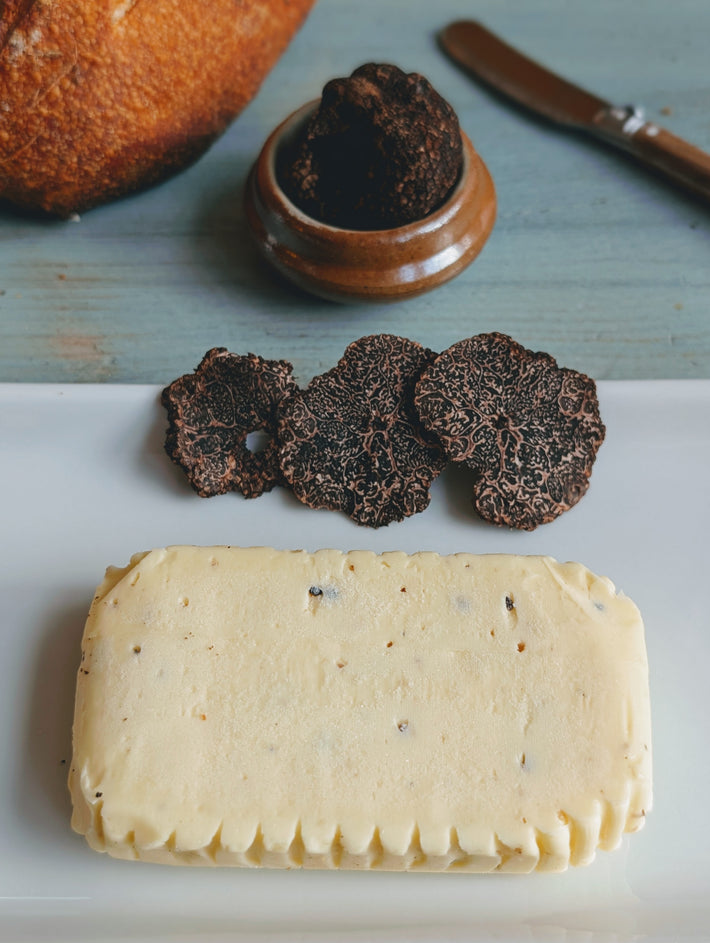 Rectangular slab of truffle butter on a white plate with sliced black truffles; jar, knife and bread blurred in background.