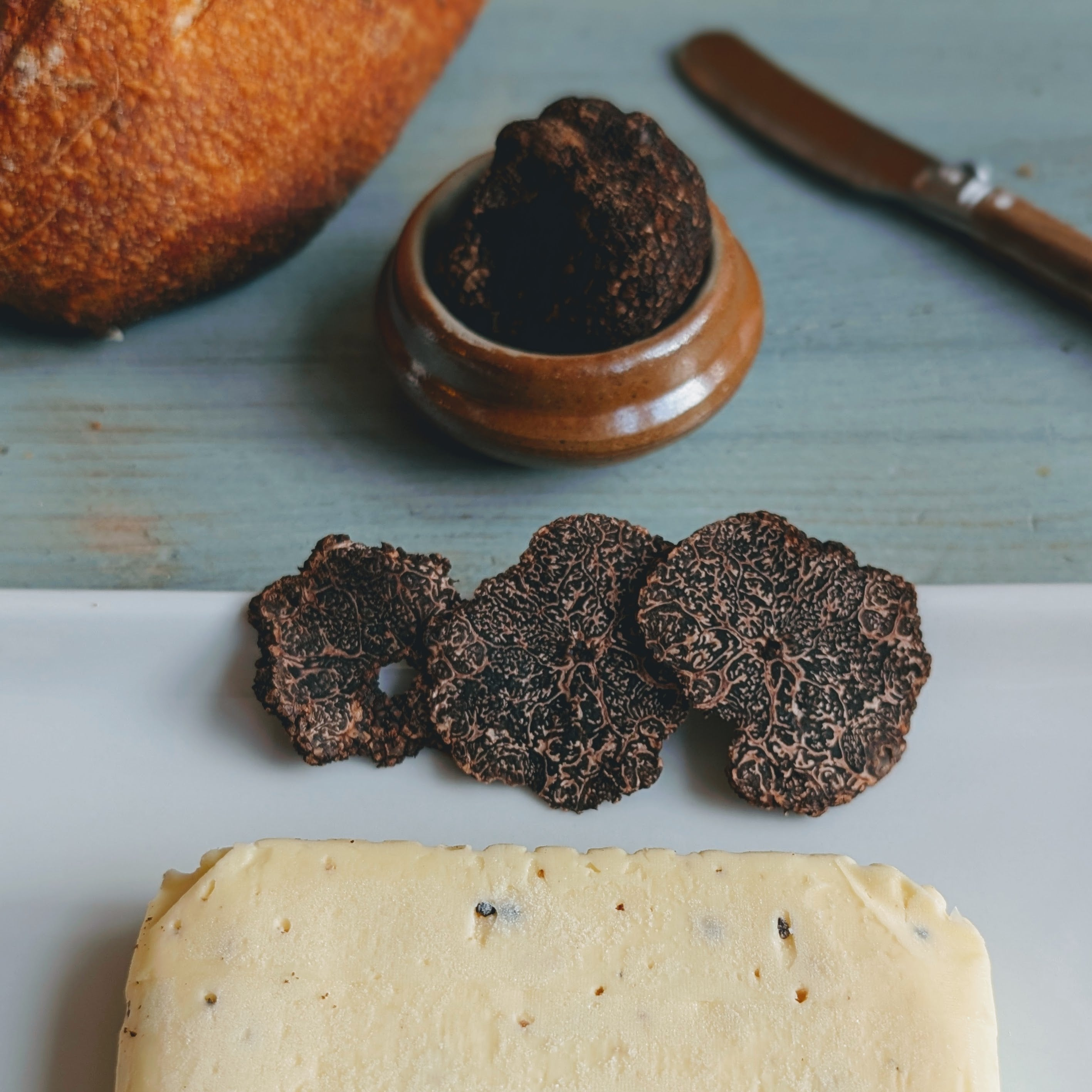 Three thin black truffle slices next to a pale truffle butter block on a plate, crusty bread and knife in background.