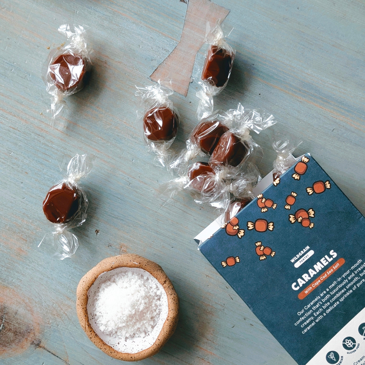 WILDGRAIN CARAMELS spilling from a box labeled 'With Cape Cod Sea Salt', beside a bowl of coarse salt.