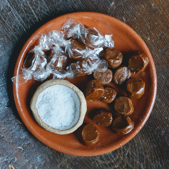Terracotta plate of wrapped and unwrapped salted caramels with a small bowl of flaky sea salt on a wooden table.
