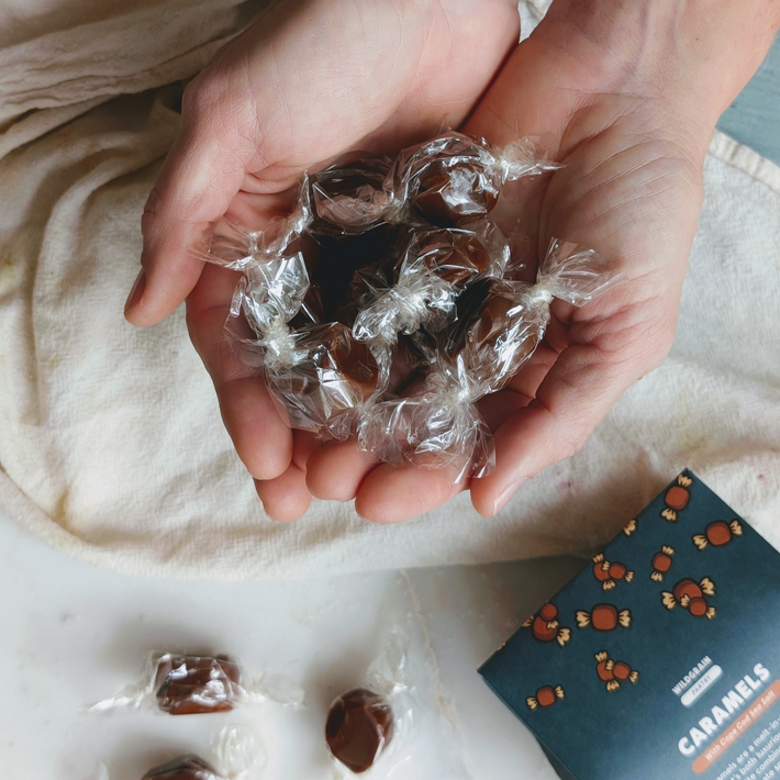 Hands cupping several individually wrapped brown caramels in clear cellophane, box labeled CARAMELS visible at lower right.
