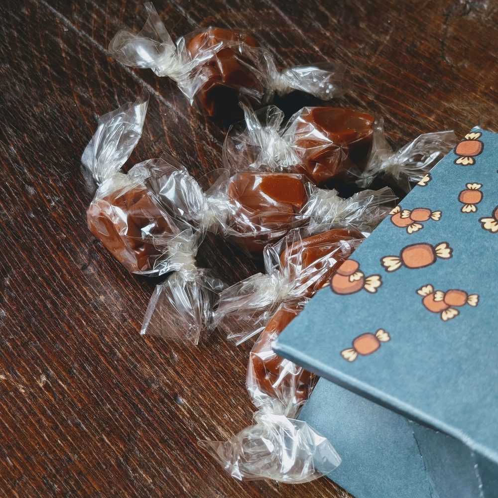 Individually wrapped brown caramels on dark wood next to a blue gift box patterned with candy illustrations.