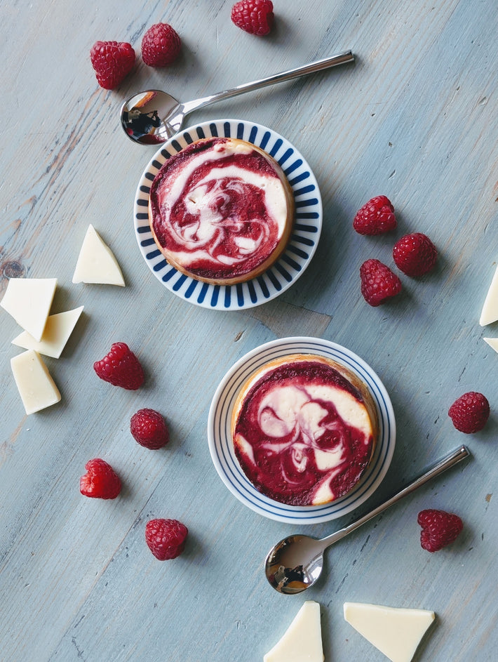Two raspberry-swirl mini cheesecakes on striped saucers with fresh raspberries, white chocolate pieces and spoons on a blue wooden table.