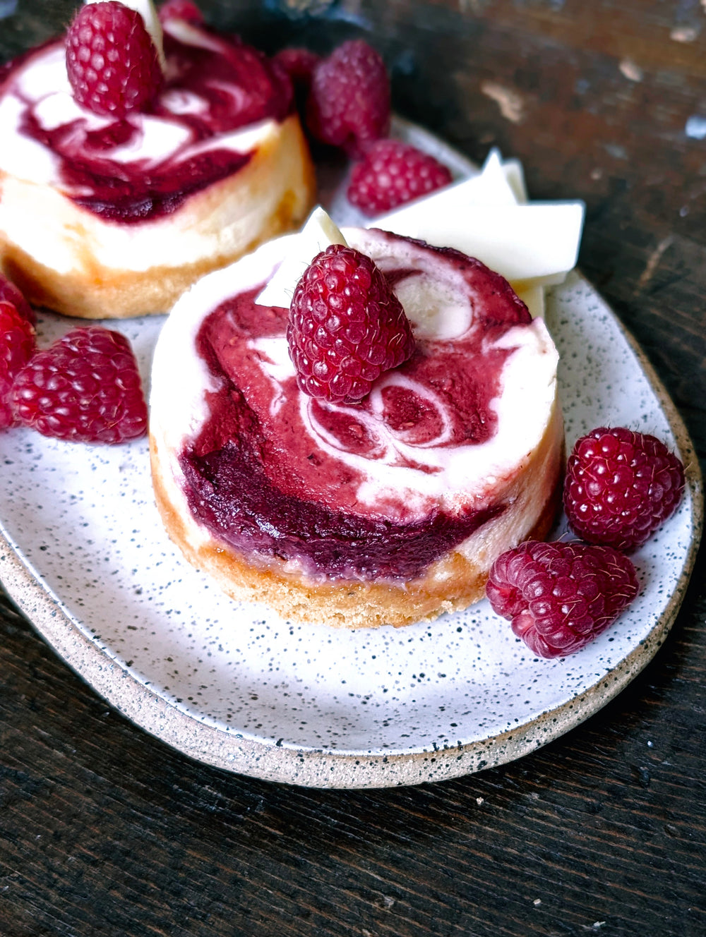 Two mini raspberry-swirl cheesecakes on a speckled plate, topped with fresh raspberries and white chocolate shards