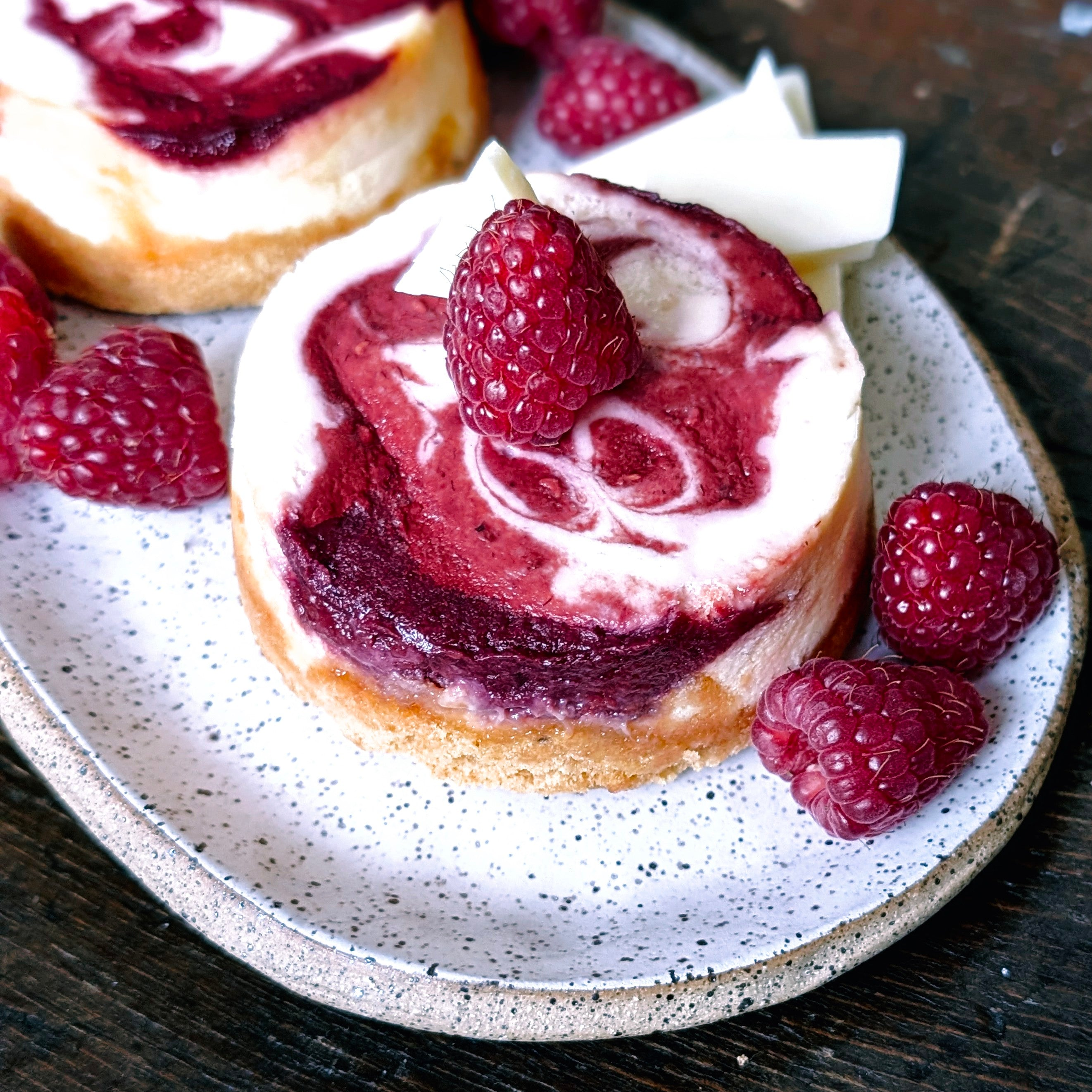 Mini raspberry swirl cheesecakes on a speckled plate topped with raspberries and white chocolate shards.