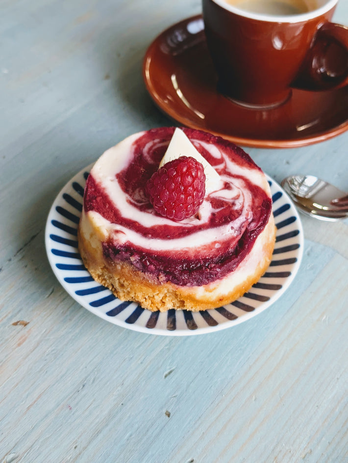 Raspberry-swirl mini cheesecake with raspberry and white chocolate triangular garnish on blue-striped plate beside coffee cup