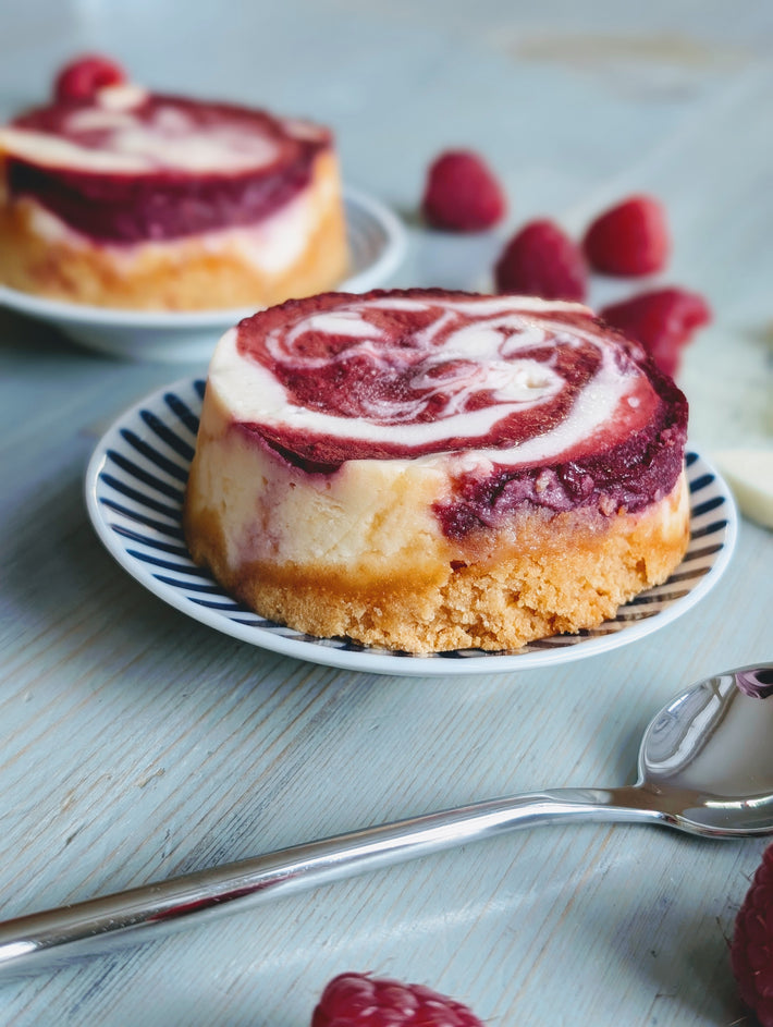 Mini raspberry-swirl cheesecake on a striped saucer with fresh raspberries and a spoon