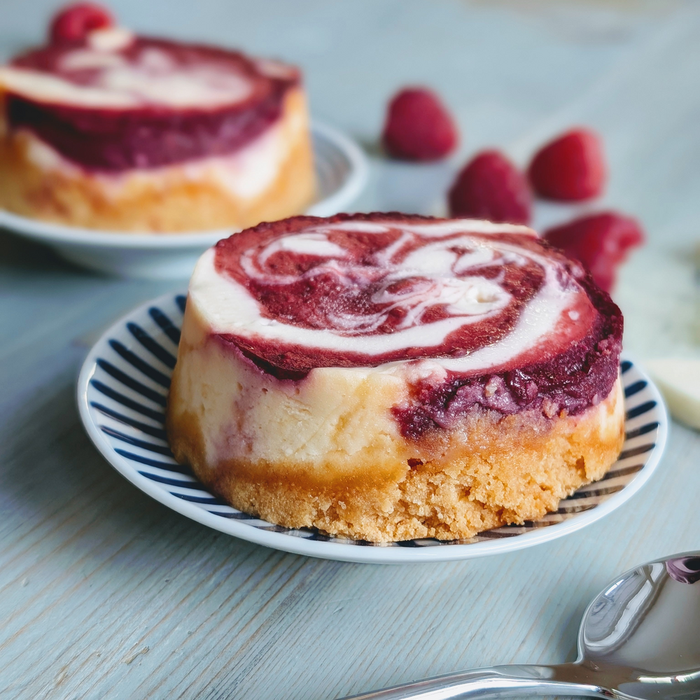 Mini raspberry swirl cheesecake on a striped saucer with fresh raspberries and a spoon