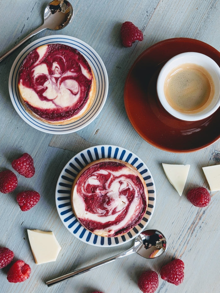 Two raspberry-swirl cheesecakes on plates with espresso, raspberries, white chocolate and spoons on gray wood.