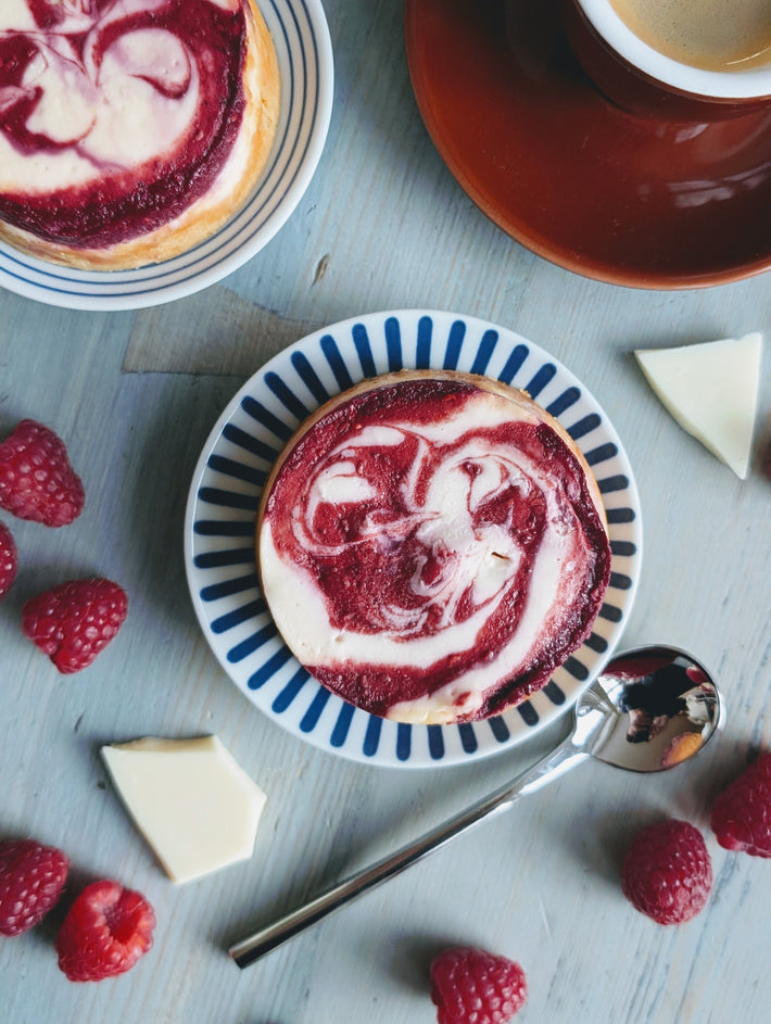 Raspberry-swirl mini cheesecake on blue-striped plate, with raspberries, white chocolate pieces, spoon and a coffee cup.
