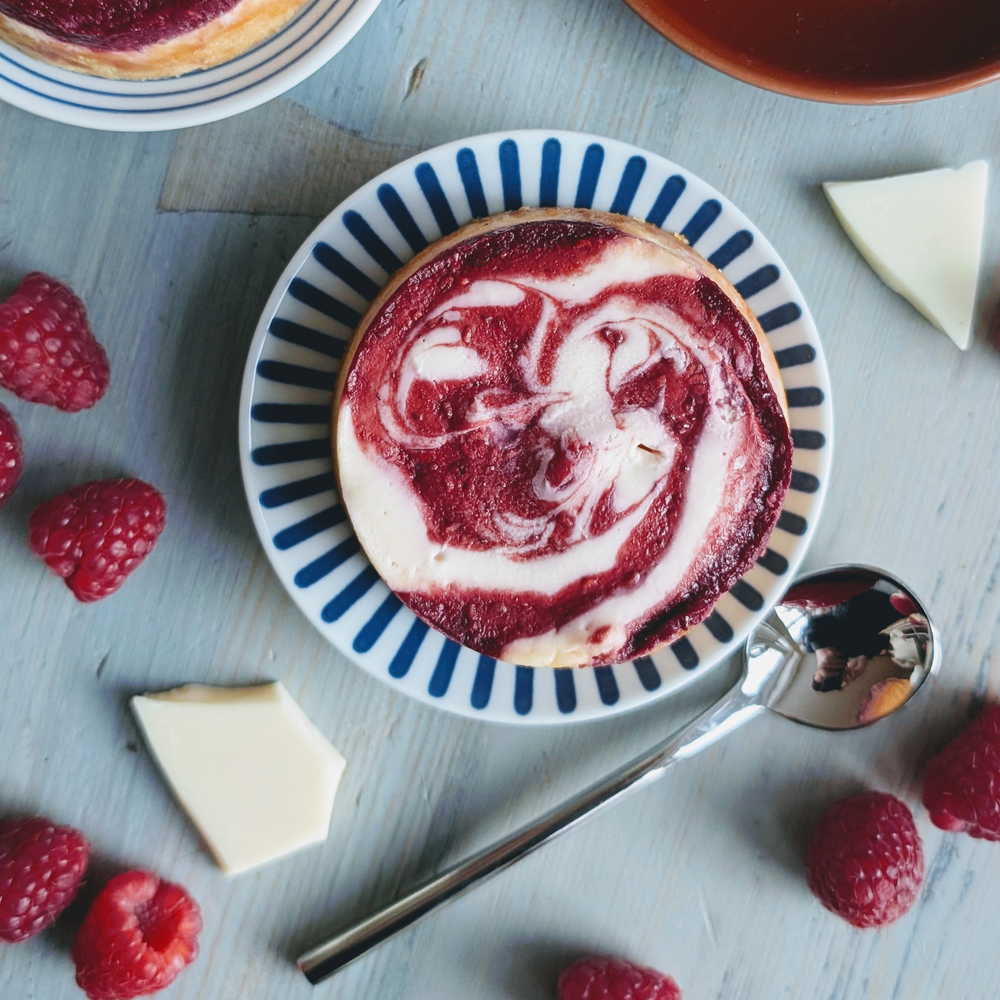 Raspberry swirl cheesecake tart on a blue-striped plate with raspberries, white chocolate pieces and a spoon.