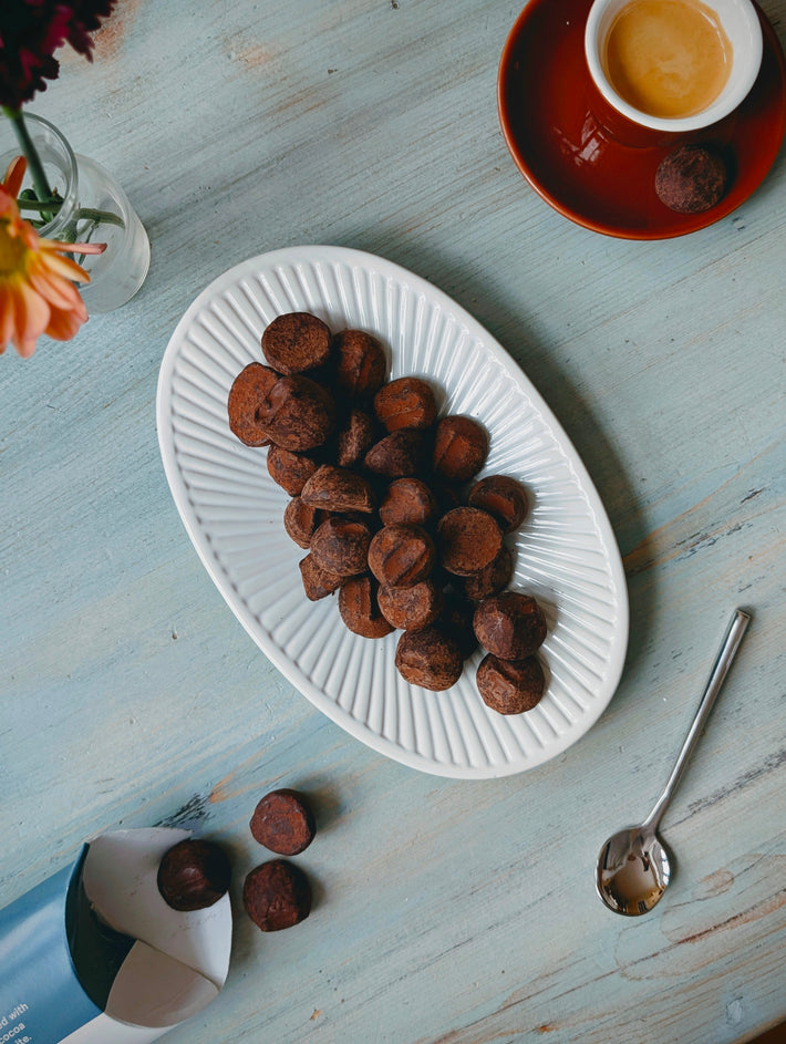Chocolate truffles on a white oval plate on pale blue wooden table, espresso cup, spoon and small vase with flowers.