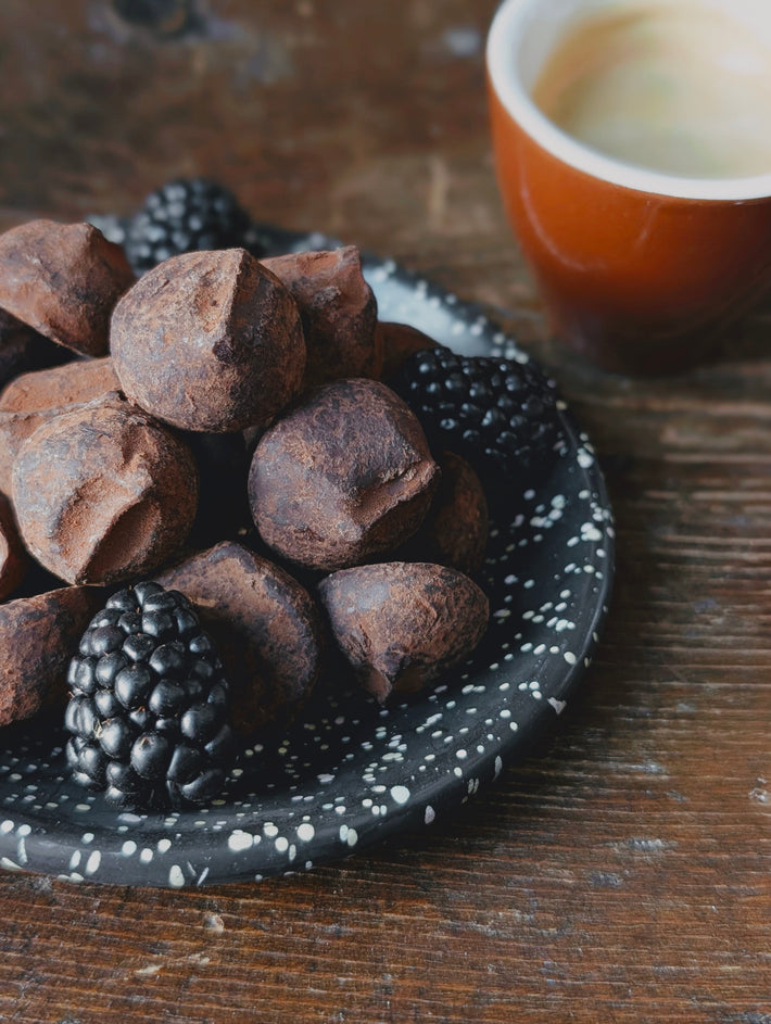 Plate of cocoa-dusted chocolate truffles and blackberries beside an espresso cup on a wooden table.