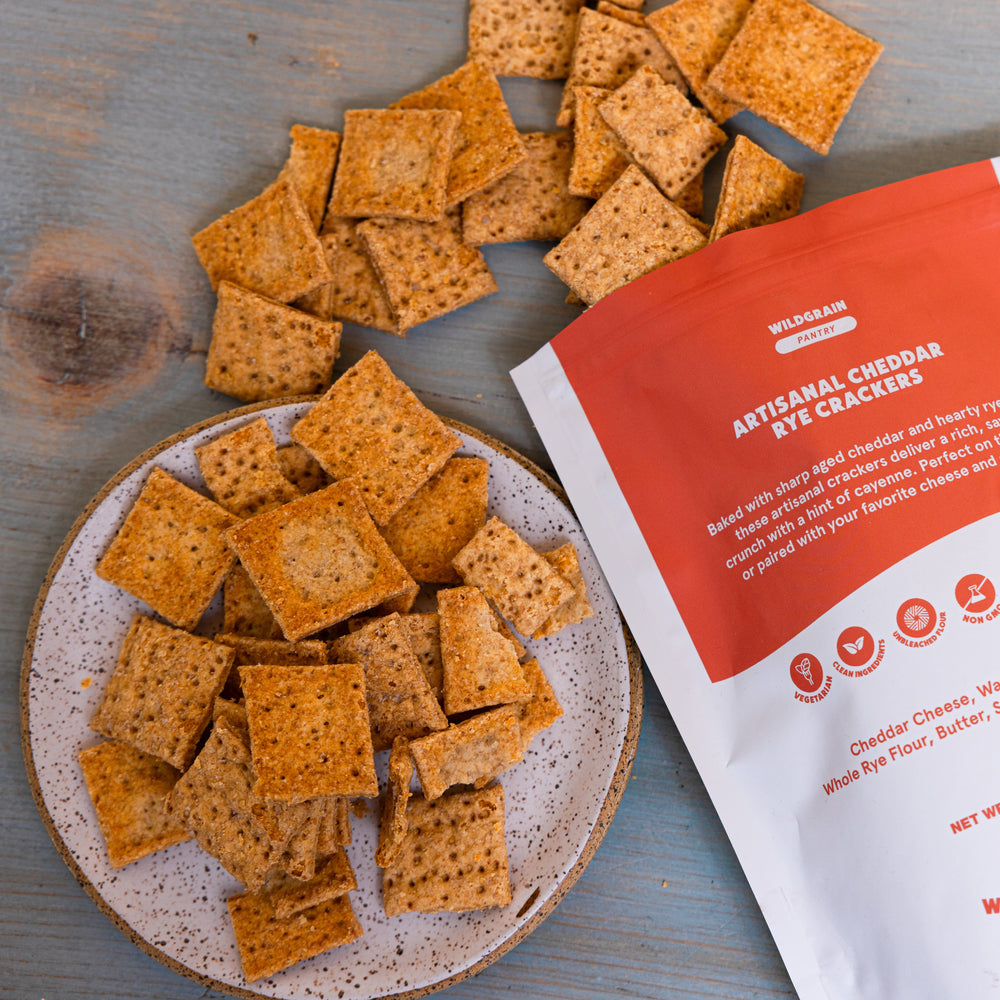 Square rye-cheddar crackers piled on a speckled plate beside a red package labeled 'ARTISAN CHEDDAR RYE CRACKERS'.