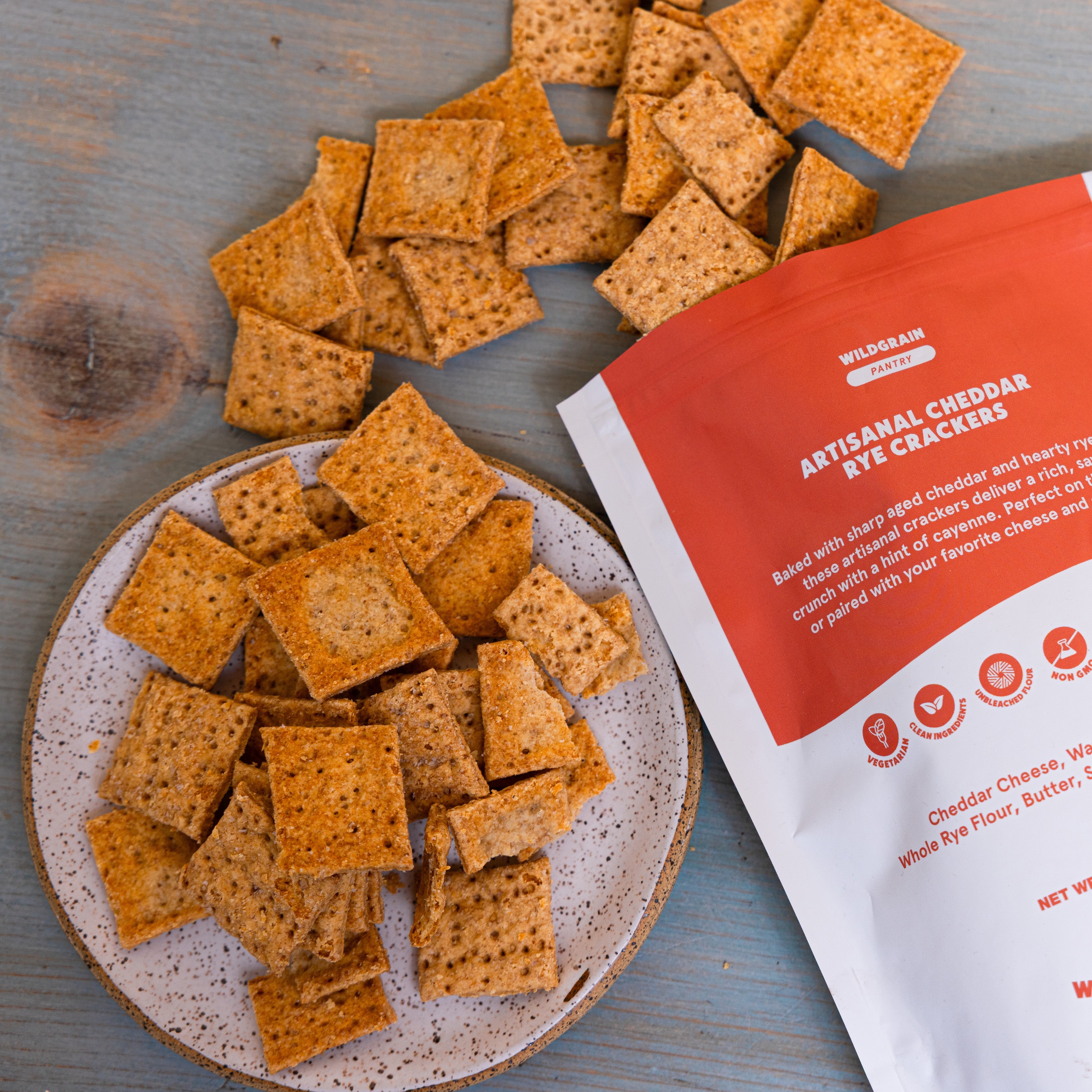 Square rye-cheddar crackers piled on a speckled plate beside a red package labeled 'ARTISAN CHEDDAR RYE CRACKERS'.