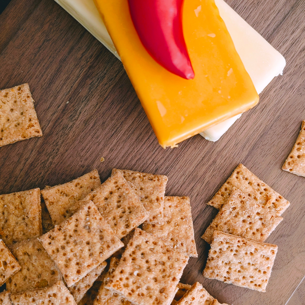 Scattered multigrain crackers on a wooden board with stacked cheddar and white cheese slices topped by a red chili pepper.