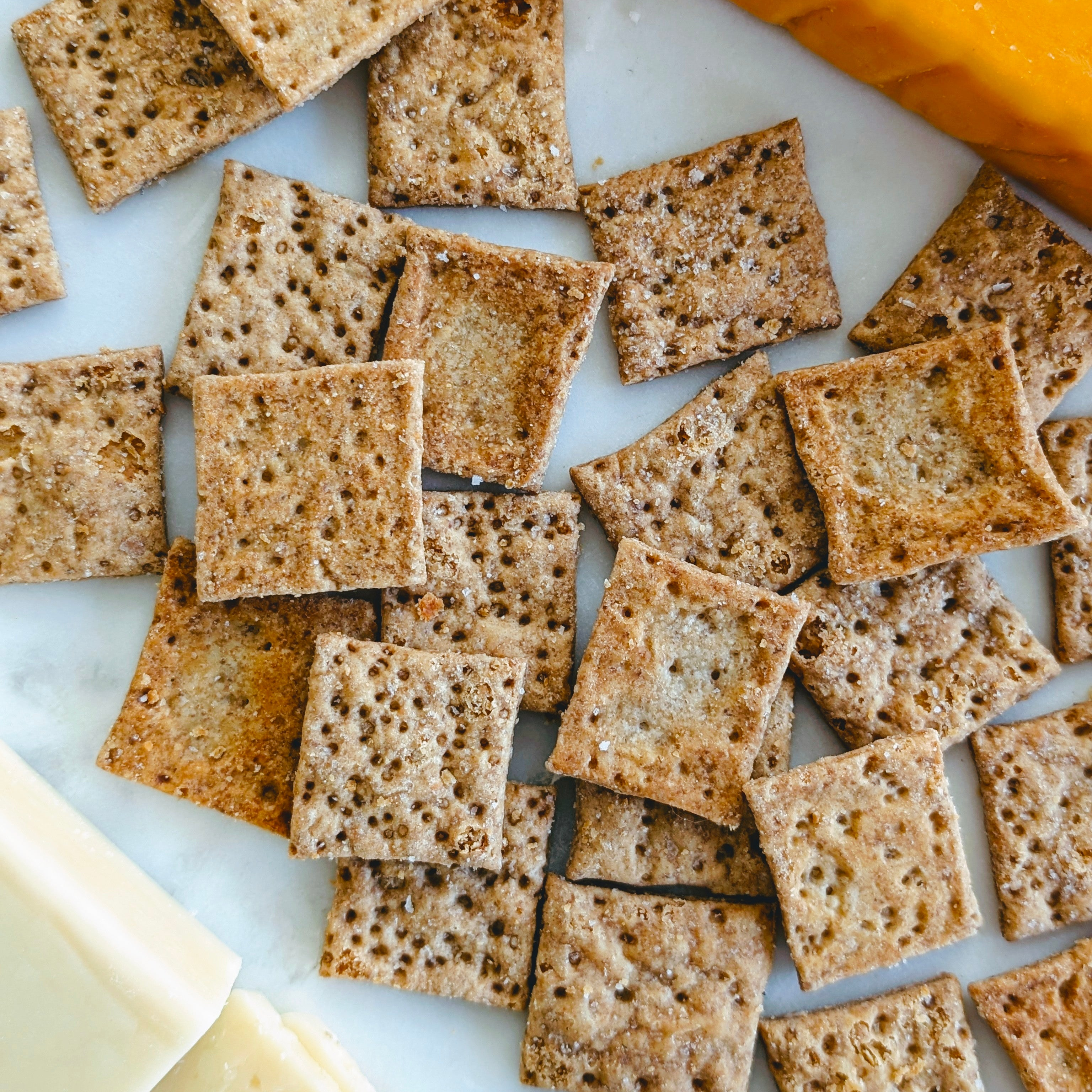 Assorted square multigrain crackers scattered on a white platter next to sliced cheese