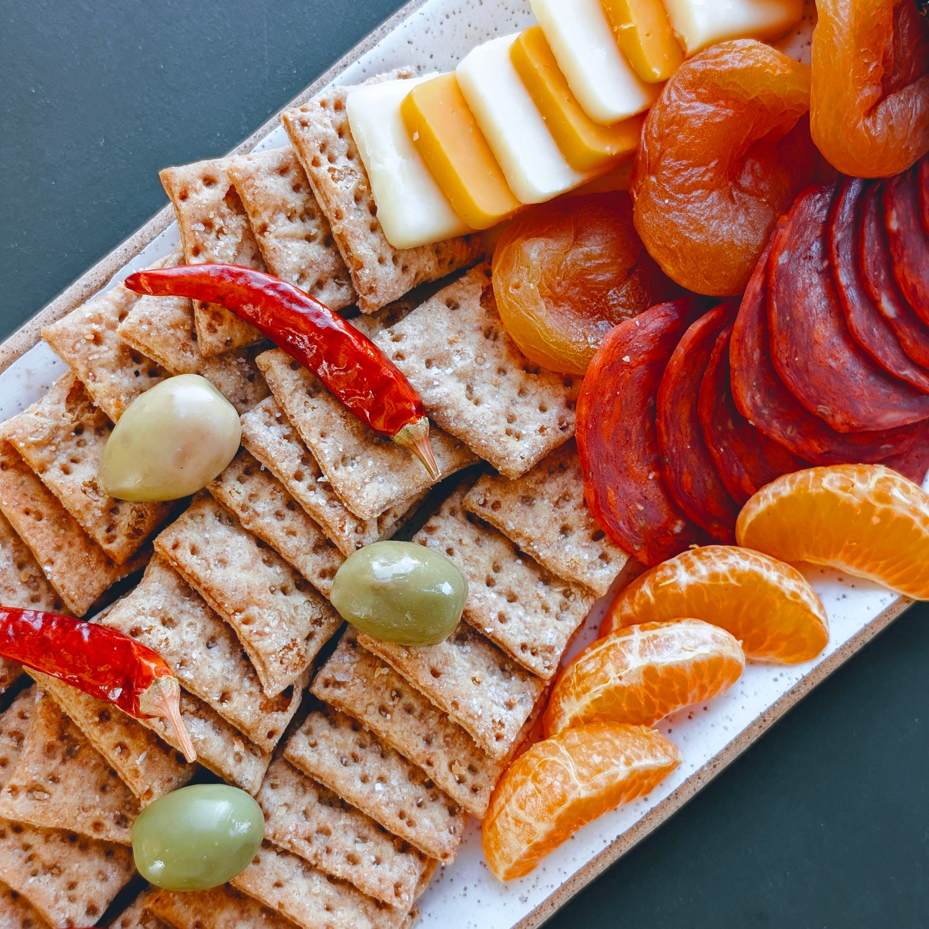 Charcuterie platter: crackers, green olives, red chilies, dried apricots, sliced cured sausage, cheese, tangerine wedges