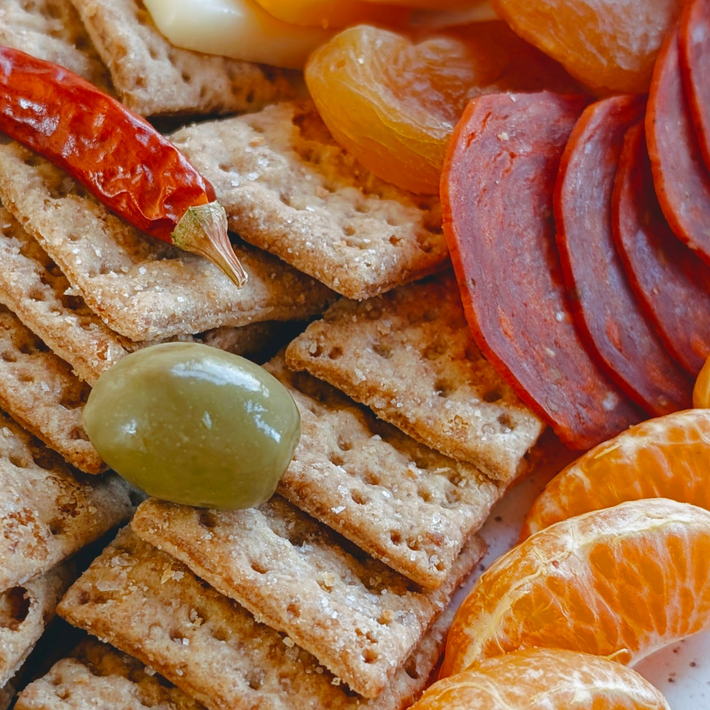 Close-up snack board with wheat crackers, green olive, folded sliced salami, dried apricots, a red chili pepper, and mandarin segments.