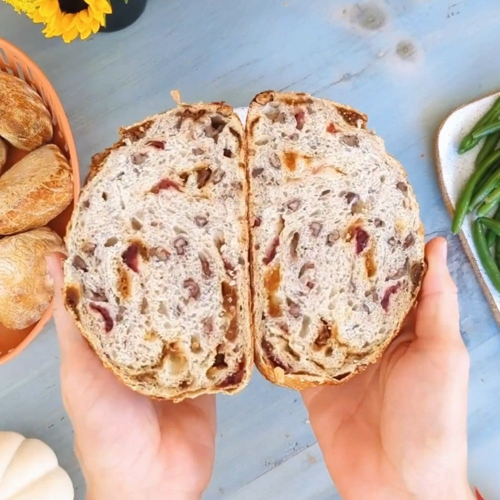 Hands holding a halved rustic loaf studded with dried fruit and nuts, shown over a table with rolls and green beans